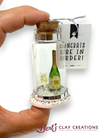 Decorative bottle with cork, miniature champagne bottle, and 'Congrats Aire In Order!' tag, held by a hand.